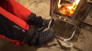 A guy is drying his shoes near a camping stove. The fire is burning and steam is coming from the shoes. Metal oven with mirror coating. Red trousers, grey mountain boots. Wet trekking shoes.