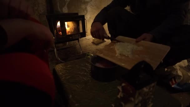 他们在不寻常的野营条件下做饭 一座古老的茅屋 篝火在营房的火炉里燃烧 蜡烛在附近 那家伙用刀把洋葱扔进一锅肉里 野营食物 — 图库视频影像