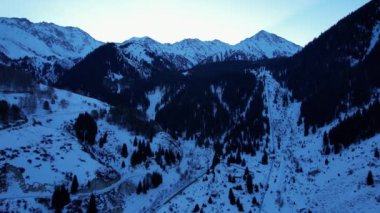Coniferous forest in snowy mountains. White clouds float across the sky. Top view from a drone. In places, the road to the mountains and buildings are visible. There is a pipe along the gorge. Sunset