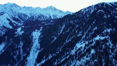 Coniferous forest in snowy mountains. White clouds float across the sky. Top view from a drone. In places, the road to the mountains and buildings are visible. There is a pipe along the gorge. Sunset