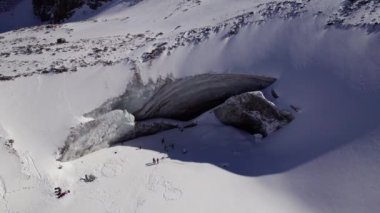 A huge frozen glacier in the mountains and a lot of tourists. Walking in the snow, taking photos with ice. Top view from a drone. Ice resembles marble. Dark light gray tones. Snowy mountains. Almaty