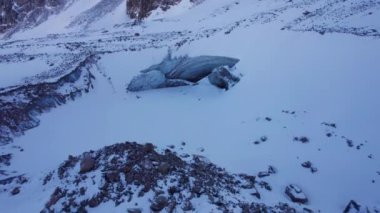 A huge ice wall from a glacier in the mountains. Everything is covered with snow and large stones. Top view from the drone on the cracks in the ice. The glacier breaks, the ice flakes off. Almaty