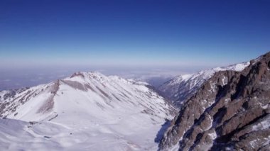 High snow-capped mountains among glaciers. Aerial view from a drone on a rocky gorge. The glacier is covered with snow and rocks. The sky is blue and the sun is shining brightly. The ice is cracking