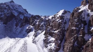 High snow-capped mountains among glaciers. Aerial view from a drone on a rocky gorge. The glacier is covered with snow and rocks. The sky is blue and the sun is shining brightly. The ice is cracking