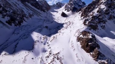 High snow-capped mountains among glaciers. Aerial view from a drone on a rocky gorge. The glacier is covered with snow and rocks. The sky is blue and the sun is shining brightly. The ice is cracking