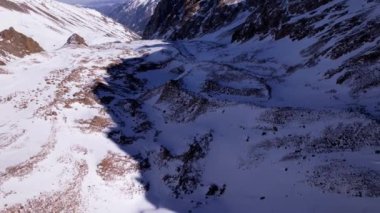 High snow-capped mountains among glaciers. Aerial view from a drone on a rocky gorge. The glacier is covered with snow and rocks. The sky is blue and the sun is shining brightly. The ice is cracking