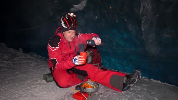 登山者在深冰洞里吃东西 从热水瓶里倒入热茶 蒸汽就来了 从煎锅里吃东西 享受食物 后面是一堵巨大的冰墙 上面有深蓝色的阴影 在山上 — 图库视频影像