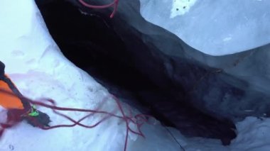 A guy pulls a climbing rope out of an ice cave. Its completely dark inside. Gray-blue ice. The climber is wearing iron cats and protection from snow. Preparing to descend into a crack in the ice