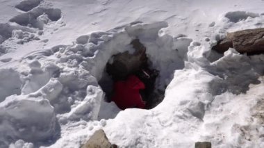 A mountaineer with a backpack descends into an ice cave. There are big rocks in the hole, white snow and ice all around. A guy in a red suit explores the entrance. Trying to get inside. Mountains