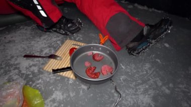 A guy cooks food on gas inside an ice cave. The climber lights the burner, cuts sausage and tomato. Breaks the eggs and fry in a frying pan. Steam from food. The ice wall is blue and the floor is gray
