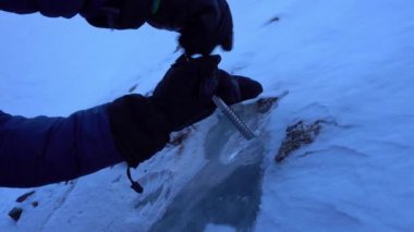 The guy twists a climbing screw into the ice. The ice drill breaks through the ice, which causes it to crack. There are frozen rocks in the glacier, white snow on top. Preparing for the descent