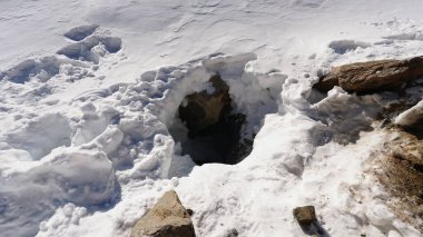 The entrance to the ice cave under the rocks and glacier. There is white snow, big stones around the hole. The tracks lead down. The entrance to the cave under the glacier. Mountains of Kazakhstan