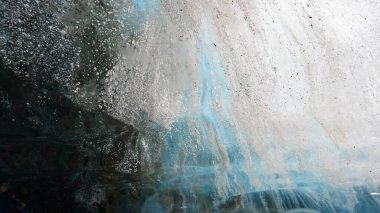 Turquoise pure color of ice inside the ice cave. Stones and icicles are visible in places. Light grains of snow on the ice walls. Frozen air bubbles in an ice wall. An ancient glacier. Color gradient