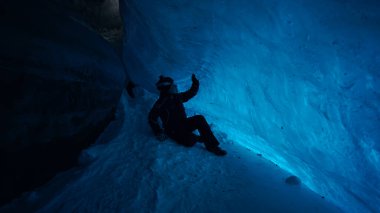 The climber made a halt inside an ice cave. The turquoise color of the ice gives a special atmosphere. The guy is looking for something in his backpack. There is snow on the icy floor. In mountains