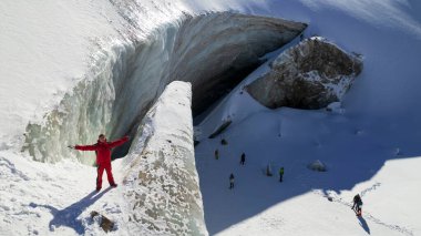 The guy in red is standing on top of the glacier. Frozen moraine lake in the mountains. Lots of snow. Aerial view from a drone of people, mountains and trails in the snow. The ice is breaking down