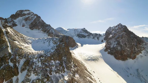 High snow-capped mountains among glaciers. Aerial view from a drone on a rocky gorge. The glacier is covered with snow and rocks. The sky is blue and the sun is shining brightly. The ice is cracking