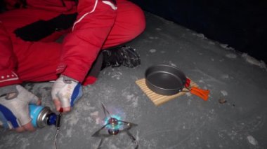 A guy cooks food on gas inside an ice cave. The climber lights the burner, cuts sausage and tomato. Breaks the eggs and fry in a frying pan. Steam from food. The ice wall is blue and the floor is gray