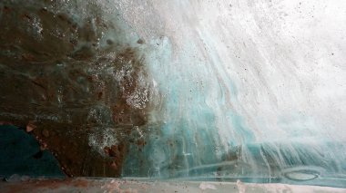 Turquoise pure color of ice inside the ice cave. Stones and icicles are visible in places. Light grains of snow on the ice walls. Frozen air bubbles in an ice wall. An ancient glacier. Color gradient