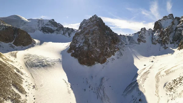 High snow-capped mountains among glaciers. Aerial view from a drone on a rocky gorge. The glacier is covered with snow and rocks. The sky is blue and the sun is shining brightly. The ice is cracking
