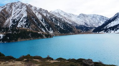 A mountain lake with blue water in winter. The suns rays are reflected from the water. Green forest and grass grow on the hills. There are stones lying. The high peaks are covered with snow. Almaty
