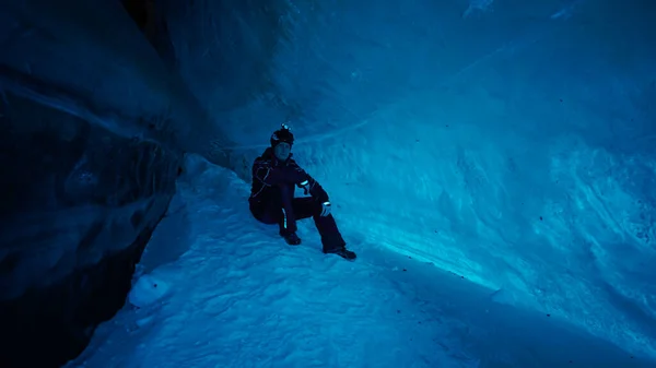 The climber made a halt inside an ice cave. The turquoise color of the ice gives a special atmosphere. The guy is looking for something in his backpack. There is snow on the icy floor. In mountains