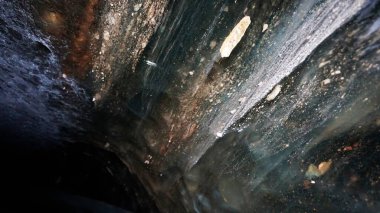 The black transparent color of ice in the cave. Small stones hang down in places. There are frozen air bubbles and grains of sand in the ice. Black-brown shade of ice. Deep in the glacier. Epic