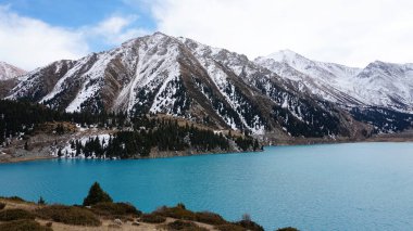 A mountain lake with blue water in winter. The suns rays are reflected from the water. Green forest and grass grow on the hills. There are stones lying. The high peaks are covered with snow. Almaty
