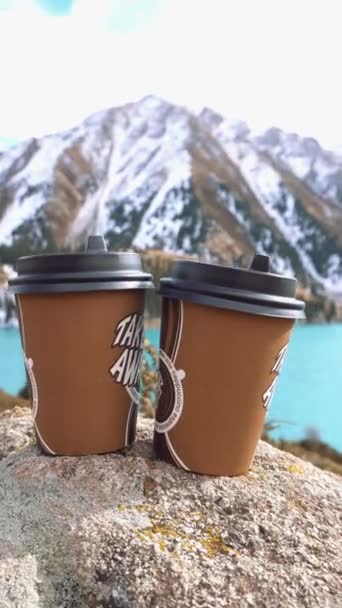 山の湖を背景にコーヒーと紙コップ 水の色は青です 丘の上に緑の森が生えている 高い山は雪で覆われている コップは石の上にある ロマンス — ストック動画