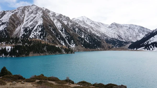 冬には青い水の山の湖 太陽光線は水から反射される 緑の森と草が丘の上に生えています 石が横になっている 高い山は雪で覆われている アルマトイ — ストック写真