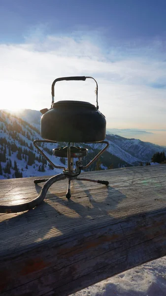 stock image The burner is burning with a blue fire with a kettle. The burner and the cylinder are on the bench. In the background there are green fir trees, mountains in white snow and a beautiful sunset