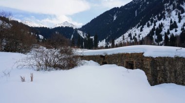 Dağlarda eski bir taş hapishane ya da kulübe. Karlı dağlar, orman ve bulutlar mavi gökyüzüne karşı. Uzun kuru çalılar karların altından dışarı bakıyor. Huş ağacı ve çam ağaçları uzaktan görülebilir..