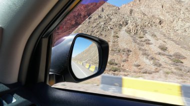 Arabanın yan aynasında yol manzarası var. Arkasında sarı çitler, kurumuş çalıları olan kayalık dağlar var. Yalan söyleyen taşlar var. Beyaz bir arabanın siyah tozlu aynası. Bir SUV. Yolculuk.