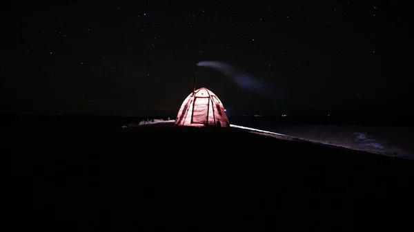 stock image A tent with a stove stands under the starry sky. Night. Bright stars. Portable stove in the tent. The light is on. Lights are visible in the distance. Grass grows on the ground and snow lies in places