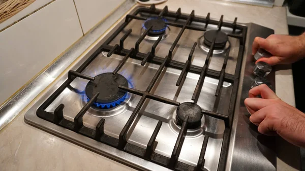 Turns off or turns on the gas on the stove. Blue flame of fire on black burners. The mans hands turn off or turn on the gas. Black grilles on a steel plate. White tiles on the walls. Cold winter