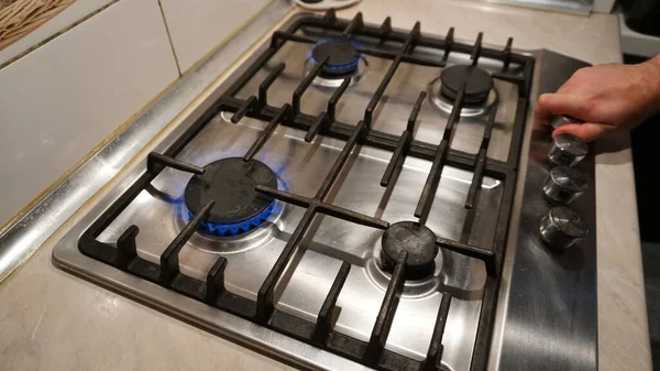 Turns off or turns on the gas on the stove. Blue flame of fire on black burners. The mans hand turns off or turns on the gas. Black grilles on a steel plate. White tiles on the walls. Cold winter