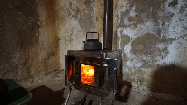 A fire is burning in a camping stove with a glass door. The process of smoldering fuel briquettes is visible. The lining of the stove made of mirror metal reflects the gray old walls of the hut.