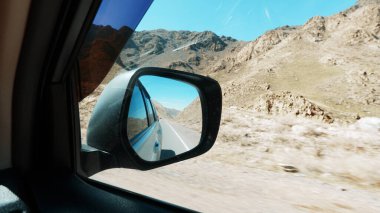 Arabanın yan aynasında yol manzarası var. Arkasında sarı çitler, kurumuş çalıları olan kayalık dağlar var. Yalan söyleyen taşlar var. Beyaz bir arabanın siyah tozlu aynası. Bir SUV. Yolculuk.