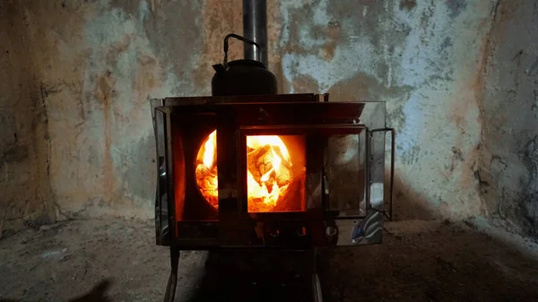 A fire is burning in a camping stove with a glass door. The process of smoldering fuel briquettes is visible. The lining of the stove made of mirror metal reflects the gray old walls of the hut.