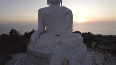 Büyük Buda, gün batımını seyrediyor. Phuket. İnsanlar heykele çıkan basamakları tırmanıyor. Adanın yeşil tepeleri her yerde. Uzakta, parlak güneş denizin üzerinde gider. Yukarıdan görüntüle
