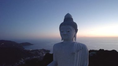 Büyük Buda, gün batımını seyrediyor. Phuket. İnsanlar heykele çıkan basamakları tırmanıyor. Adanın yeşil tepeleri her yerde. Uzakta, parlak güneş denizin üzerinde gider. Yukarıdan görüntüle