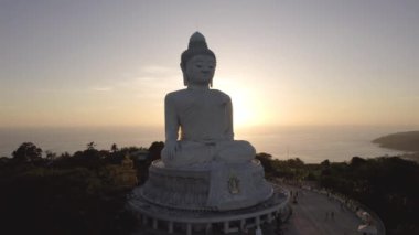 Büyük Buda, gün batımını seyrediyor. Phuket. İnsanlar heykele çıkan basamakları tırmanıyor. Adanın yeşil tepeleri her yerde. Uzakta, parlak güneş denizin üzerinde gider. Yukarıdan görüntüle