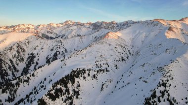 Yüksek karlı dağlarda destansı kırmızı-turuncu gün batımı. Uzun kozalaklı ağaçlar var, tepelerde kar var. Yüksek zirveler. Güneş ışınları bulutların üzerine düşer ve sis vadiden aşağı iner. Almaty