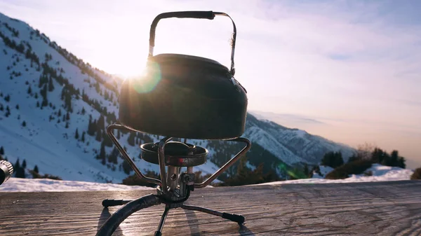 stock image The burner is burning with a blue fire with a kettle. The burner and the cylinder are on the bench. In the background there are green fir trees, mountains in white snow and a beautiful sunset