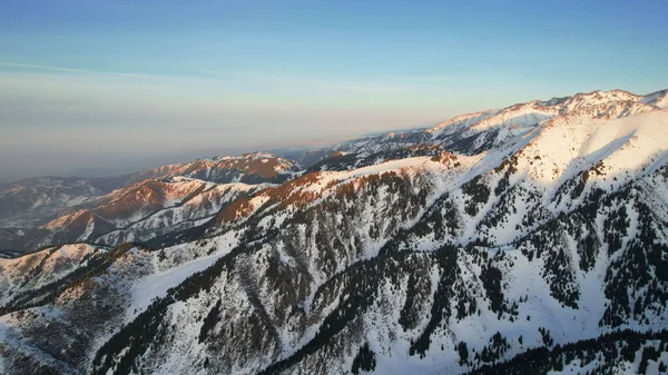 stock image Epic red-orange sunset in high snowy mountains. There are tall coniferous trees, there is snow on the hills. High peaks. The suns rays fall on the clouds and fog that is lower down the gorge. Almaty