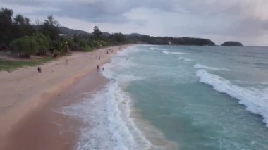 Deniz manzaralı ve bulutlu güzel bir gün batımı. Kumlu bir sahil ve su üzerindeki bir dronun üst görüntüsü. Turistler rahatlar ve yüzerler. Phuket Adası, Tayland 'ın yeşil tepeleri. Evler ve yüksek palmiye ağaçları