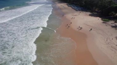 Okyanus manzaralı, dalgalı ve plajlı. Açık sarı kum suyla ıslaktır. Yeşil palmiye ağaçları. İnsanlar yürüyor. Güneş pansiyonları var. Binalar ve yeşil tepeler görülebilir. Phuket