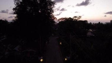 Gün batımında palmiye ağaçlarının arasından otele giden yol. Bakımlı çimenler ve çalılar. Çiçek saksılarında sarı ışık. Düşük seviyeli evler var. Uzakta, turuncu bir gün batımı ve bulutlu bir deniz.