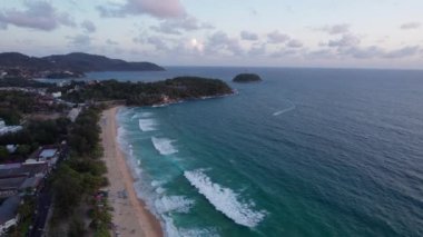 Deniz üzerinde bulutlu kırmızı bir günbatımı manzarası. Yeşil ada Phuket. Kumsalda sarı kum. İnsanlar dinleniyor. Kıyıya yakın dalgalar. Oteller var. Suyun üzerindeki turuncu güneş ışınları
