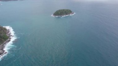 Okyanusun ortasındaki yeşil bir adanın en üst görüntüsü. Bulutlardan koyu yeşil su. Gün batımı. Dalgalar adanın kayalarına vuruyor. Dalgalı deniz. İnsansız hava aracının görüntüsü. Karon Sahili, Phuket