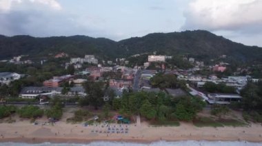 Deniz manzaralı ve bulutlu güzel bir gün batımı. Kumlu bir sahil ve su üzerindeki bir dronun üst görüntüsü. Turistler rahatlar ve yüzerler. Phuket Adası, Tayland 'ın yeşil tepeleri. Evler ve yüksek palmiye ağaçları